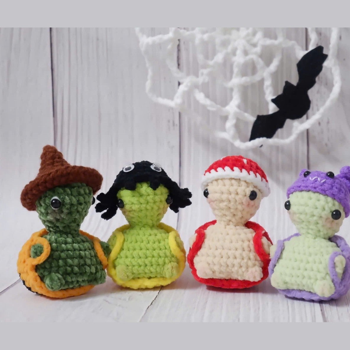Four small crocheted amigurumi figures resembling Halloween characters with witch hat, bat wings, Santa hat, and devil horns.
