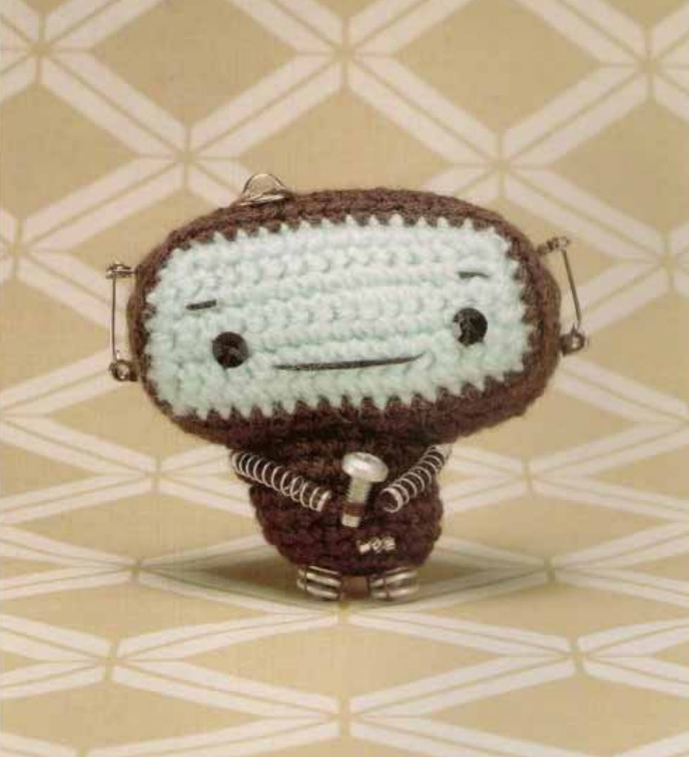 Small brown and cream crochet amigurumi robot with stitched eyes and mouth on patterned beige background.