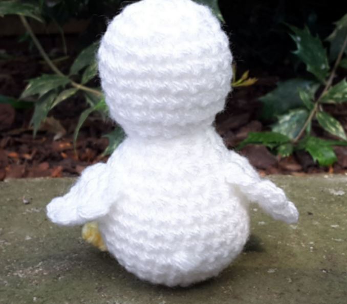 White crochet amigurumi duckling with round body and small wings sitting outdoors on stone surface.