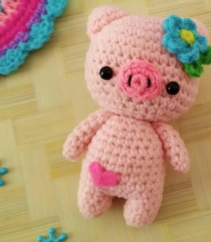 Pink crocheted amigurumi pig with blue flower on head and small heart detail on leg.