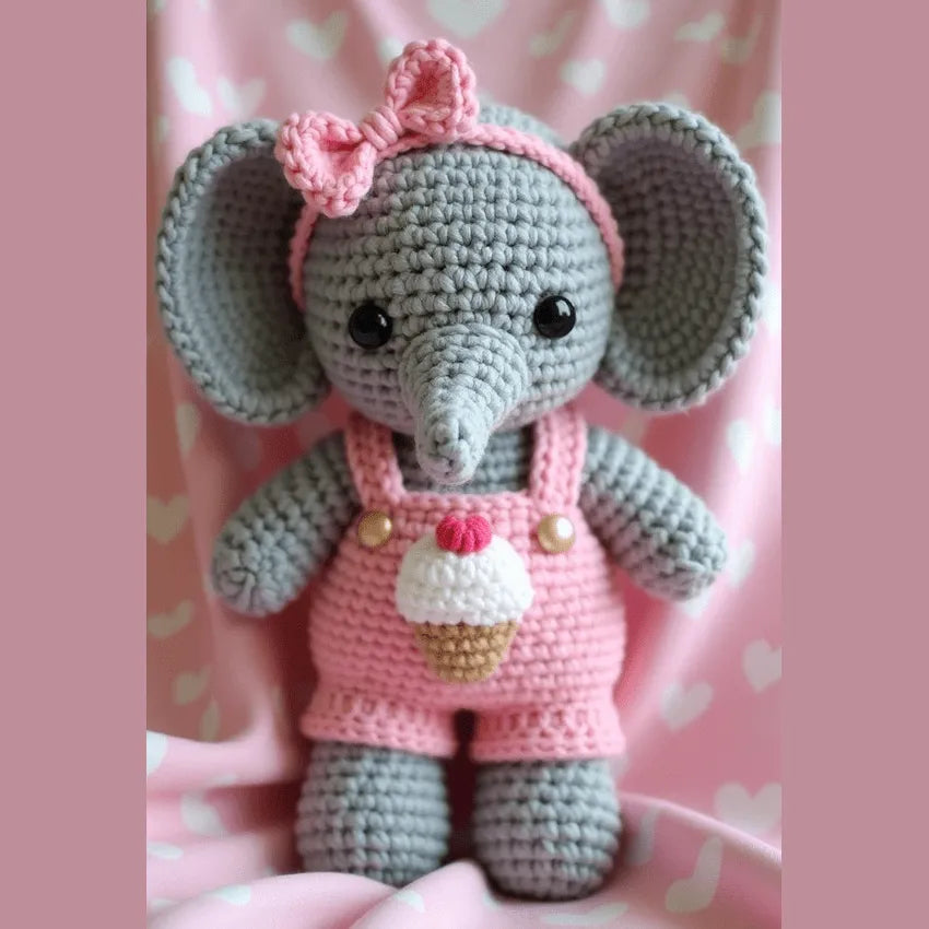 Gray crocheted amigurumi elephant wearing pink overalls with cupcake design and matching bow headband against pink background.