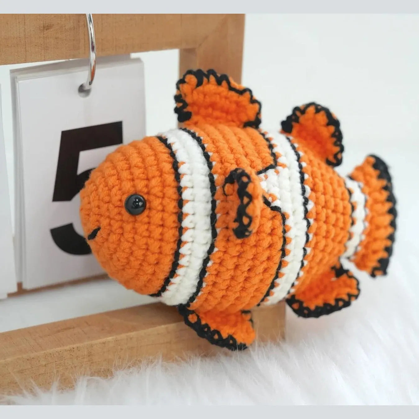 Orange and white striped amigurumi clownfish with black accents next to a wooden calendar block displaying the number 5.
