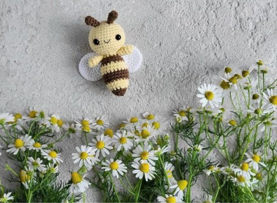 Small crocheted amigurumi bee with brown and yellow stripes, white wings, and black eyes on gray background near white.