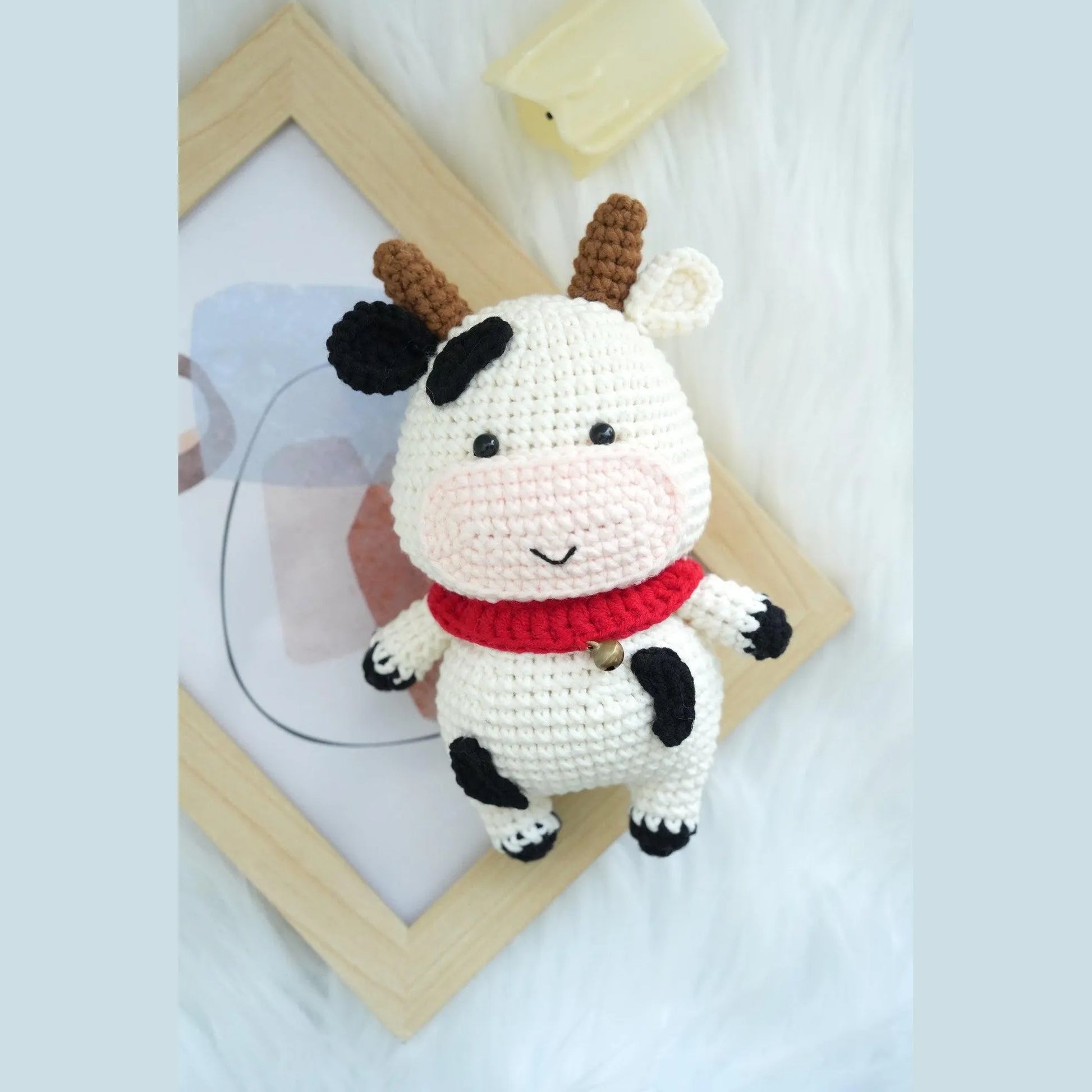 Small white and black spotted crochet amigurumi cow with red scarf on white fuzzy surface.