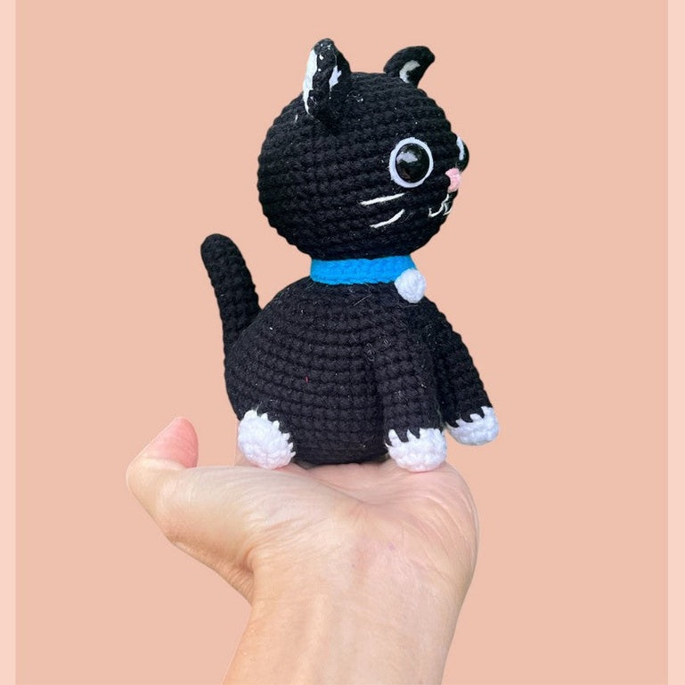 Black crocheted amigurumi cat with blue collar and white paws held in hand against peach background.