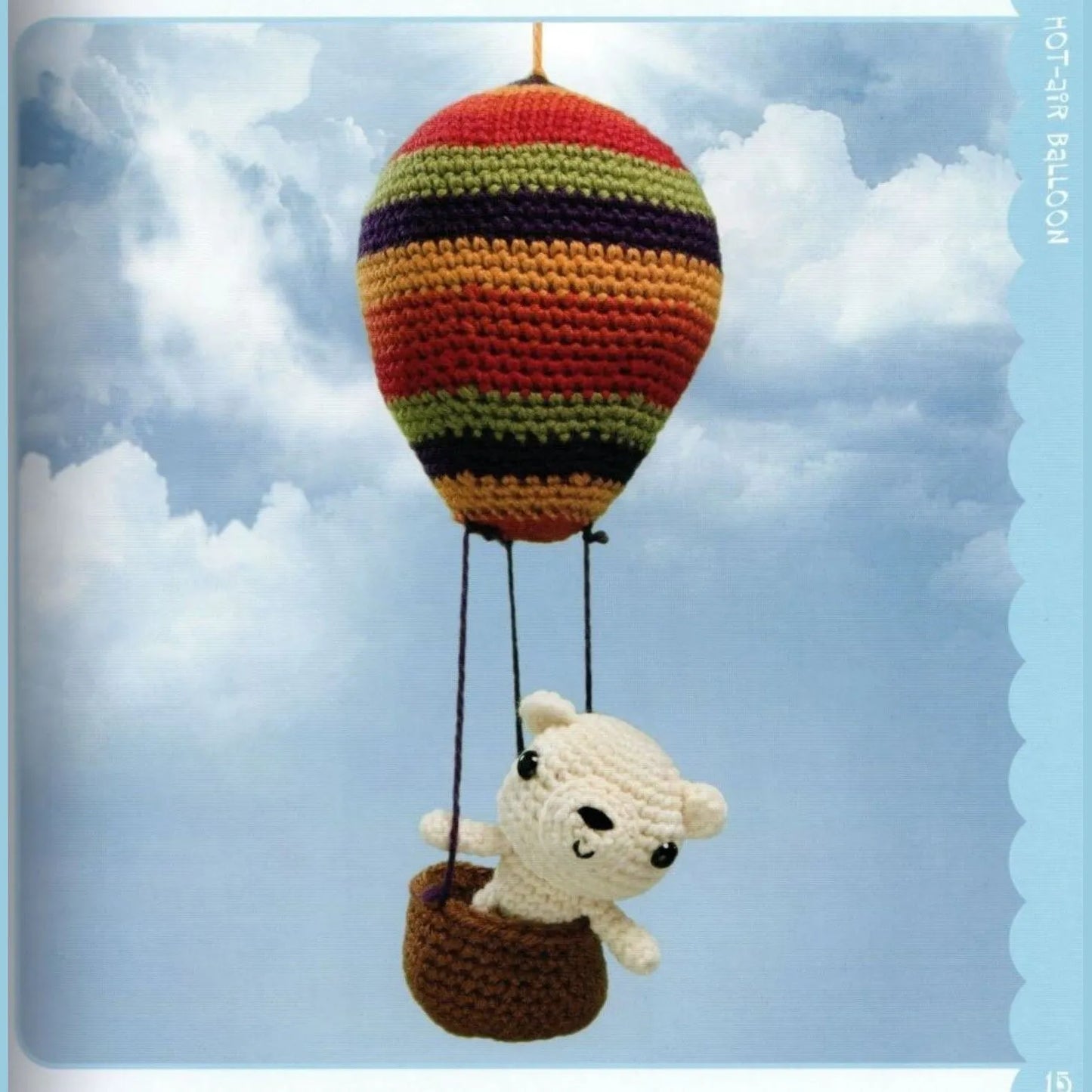 Crocheted amigurumi bear in brown basket hot air balloon with red, yellow, and black stripes against blue sky.
