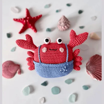 Red amigurumi crab wearing blue shorts with seashells and starfish background.