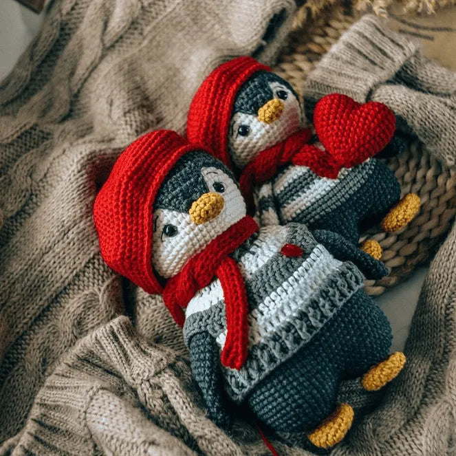 Two handmade crochet amigurumi penguins wearing red hats and scarves, one holding a red heart, on a knitted beige background.