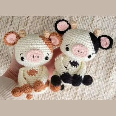 Two small crocheted amigurumi animals, a pig and a cow, with round eyes and pink snouts, held in hand on a textured knit.