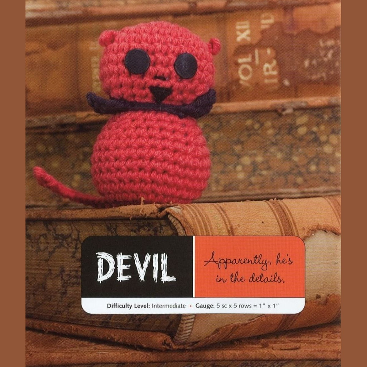 Red crocheted amigurumi devil with black eyes, nose, and mustache sitting on a vintage book.