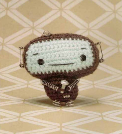 Small brown and cream crochet amigurumi robot with stitched eyes and mouth on patterned beige background.