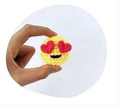 Yellow crochet amigurumi emoji with heart-shaped eyes and smiling mouth, held by hand.