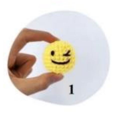 Small yellow amigurumi emoji with a winking smiley face held between fingers against a white background.