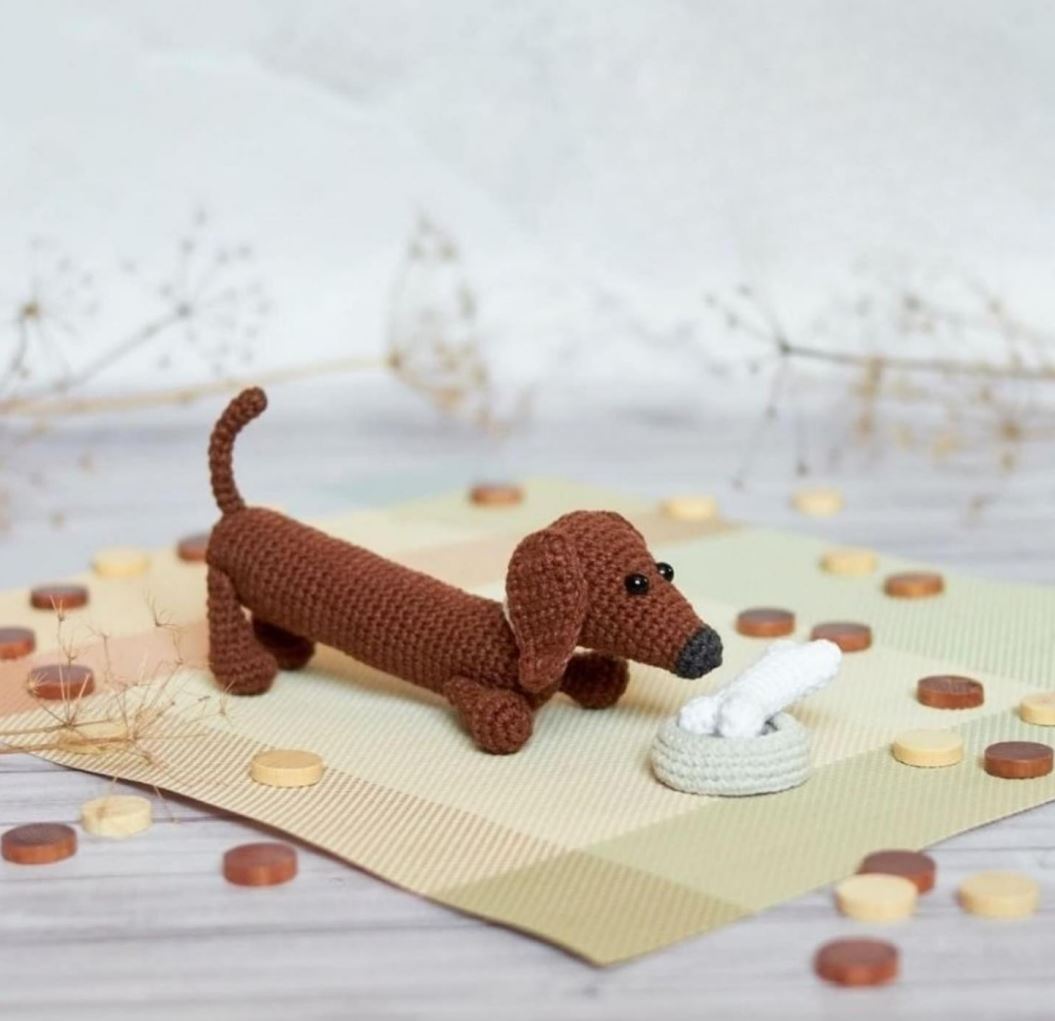 Brown crocheted dachshund amigurumi with black nose and eyes next to a small white bone on a beige mat.