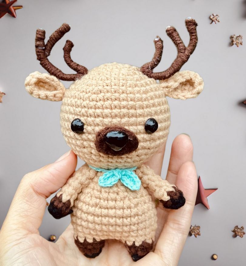Beige crocheted deer amigurumi with dark brown antlers, hooves, nose, and teal bowtie.
