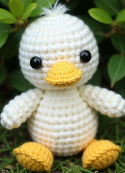 Small white crocheted amigurumi duck with black eyes, yellow beak, and feet sitting on grass.