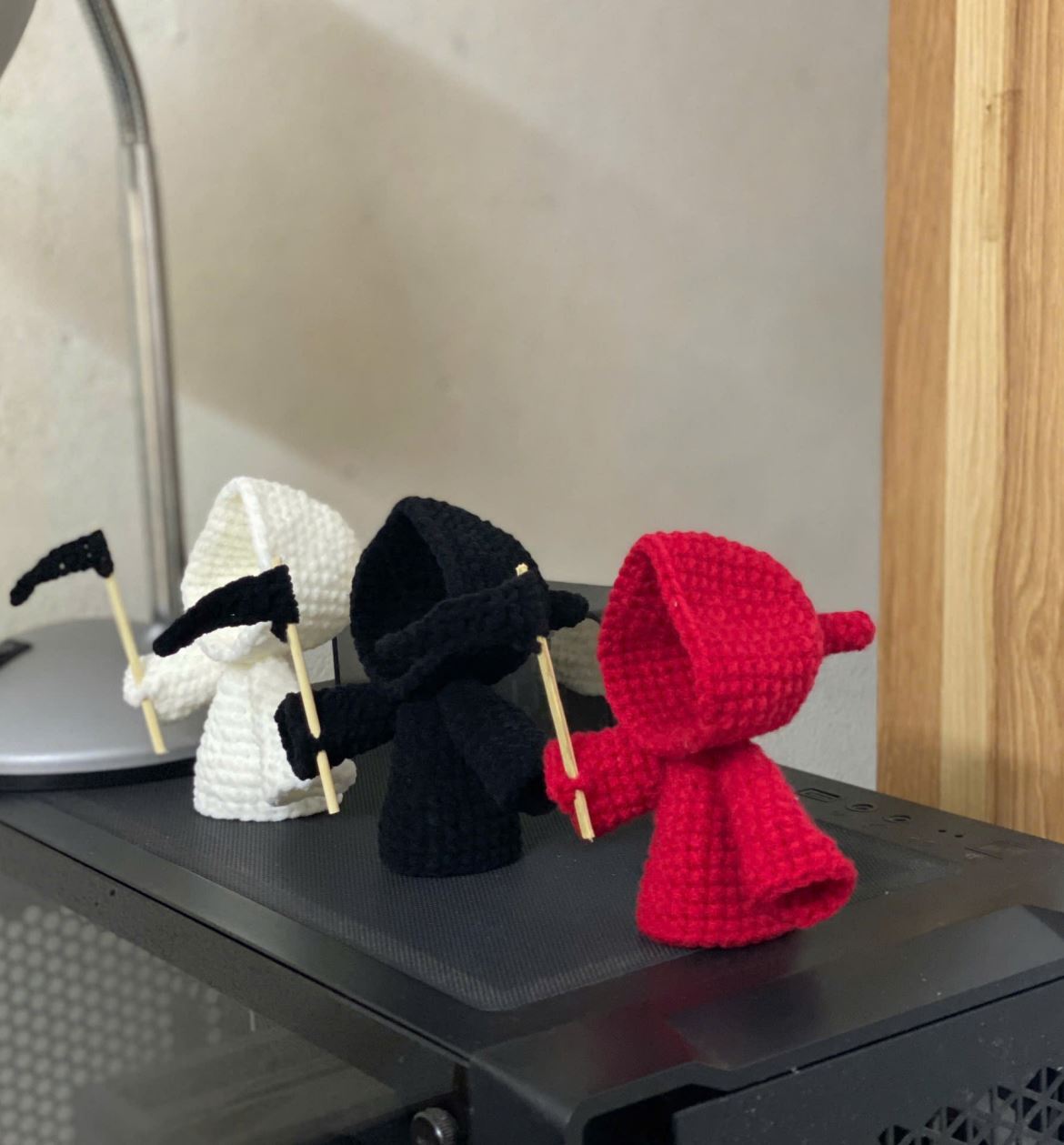 Red, black, and white crochet grim reaper amigurumi figurines holding mini scythes on a black surface.