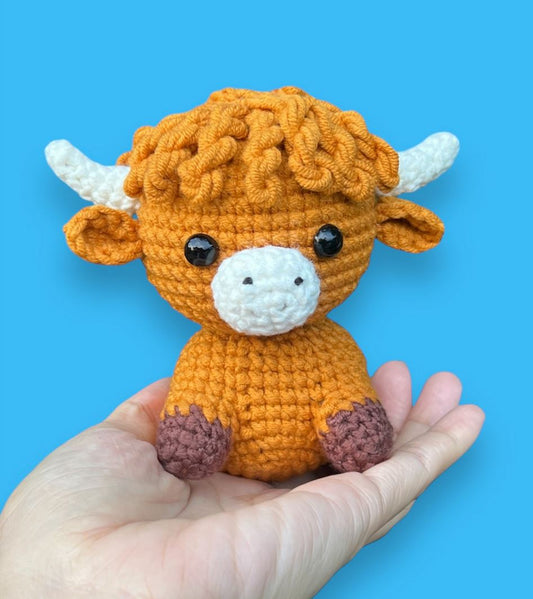 Handheld fluffy orange Highland cow amigurumi with white horns and brown hooves against blue background.