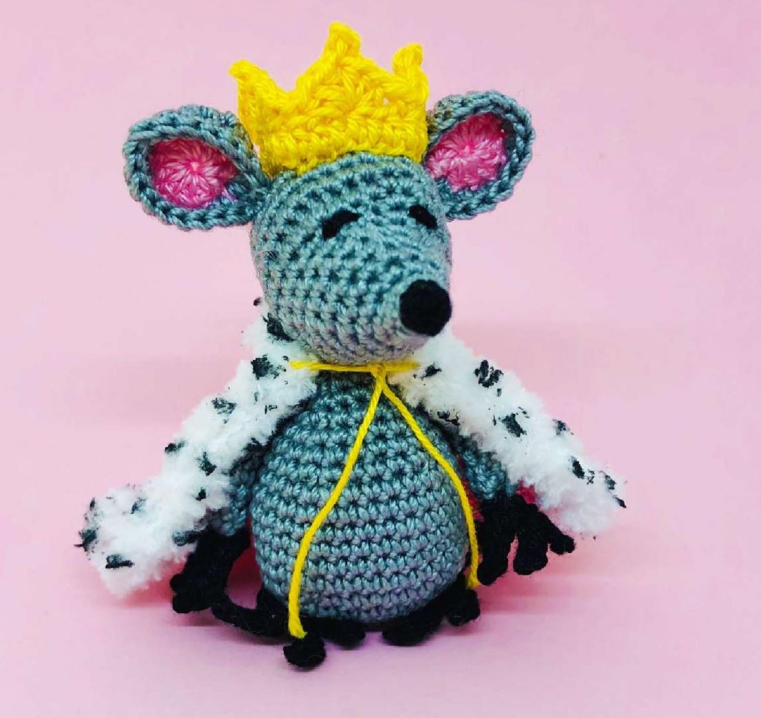 Crochet Mouse King amigurumi with yellow crown and royal fur cape against pink background.
