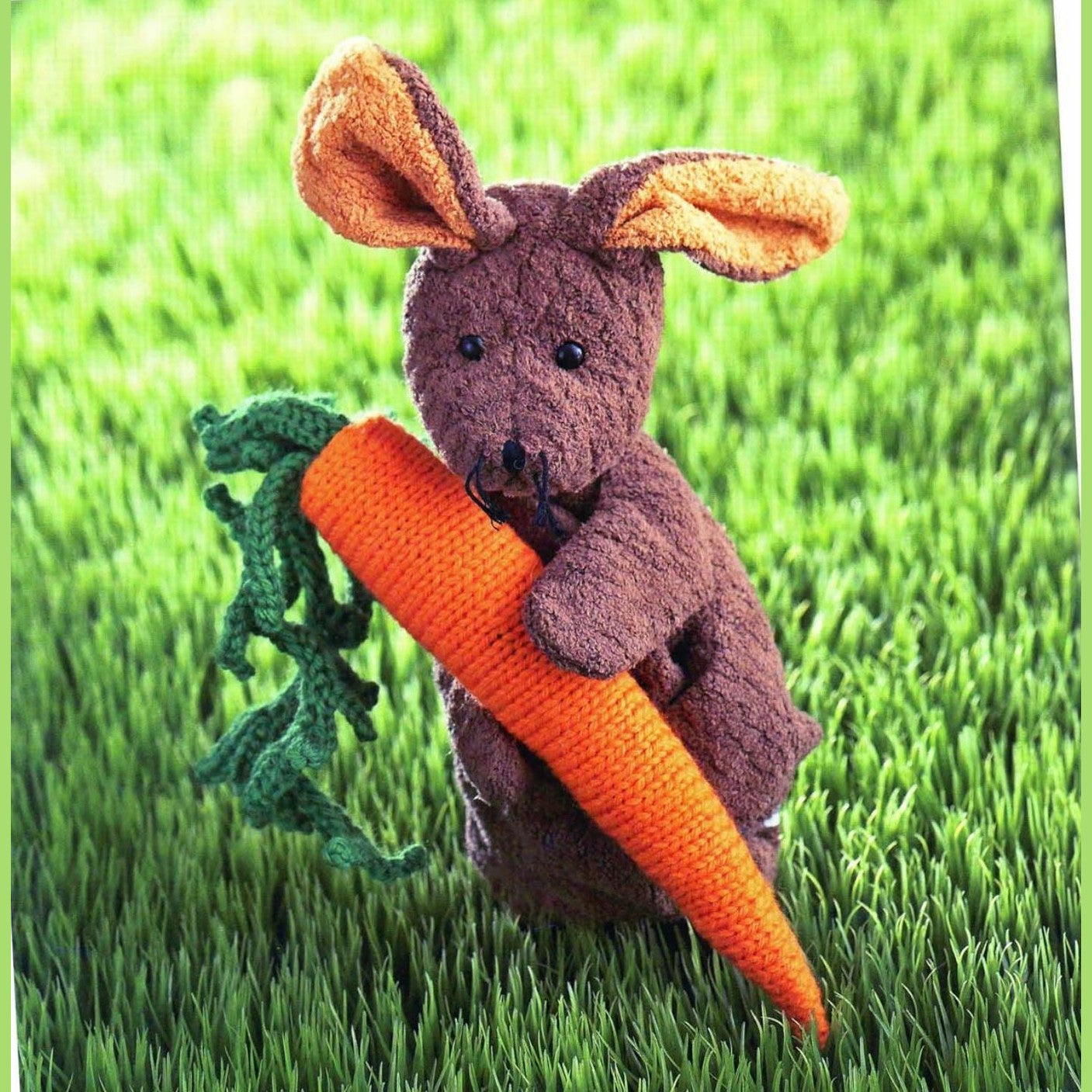 Brown knitted amigurumi bunny holding bright orange knitted carrot on green grass.