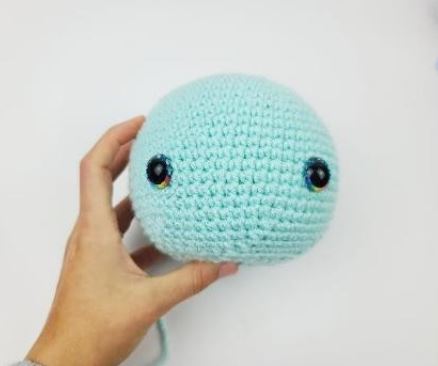 Light blue crocheted amigurumi head with black and orange eyes held in hand on white background.
