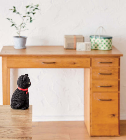 Black crocheted amigurumi cat with red collar sitting on wooden chair beside wooden desk.