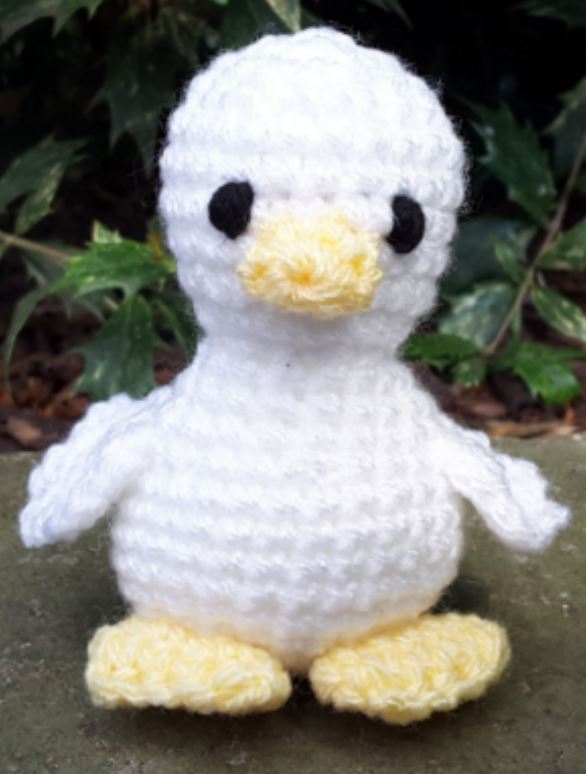 White and yellow baby duck amigurumi with black eyes sitting outdoors.