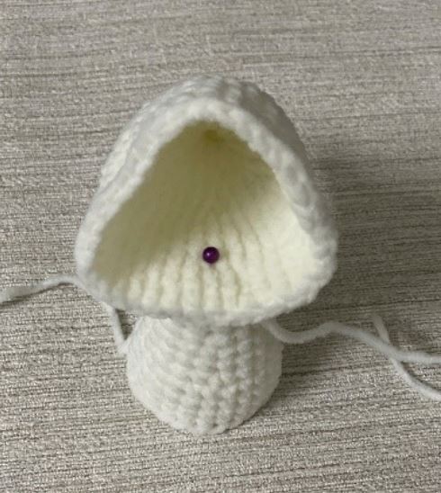 White crocheted amigurumi hood piece on beige fabric background with purple pin.