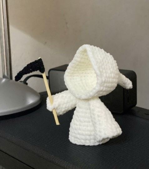 White crocheted grim reaper amigurumi with hood holding small black scythe on wooden stick.