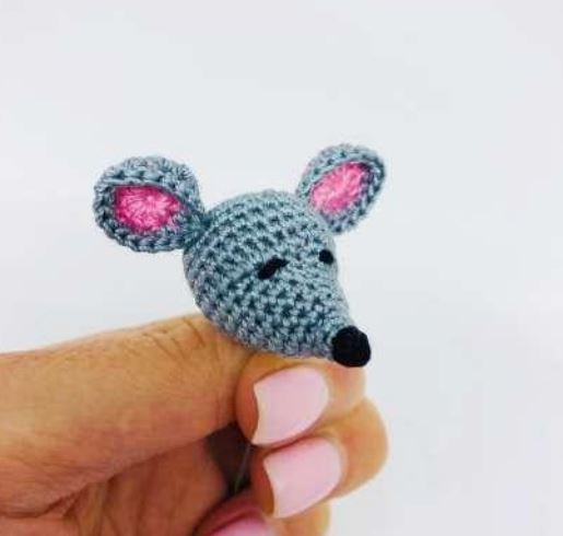 Small blue and pink crochet amigurumi mouse head with black nose held between fingers.