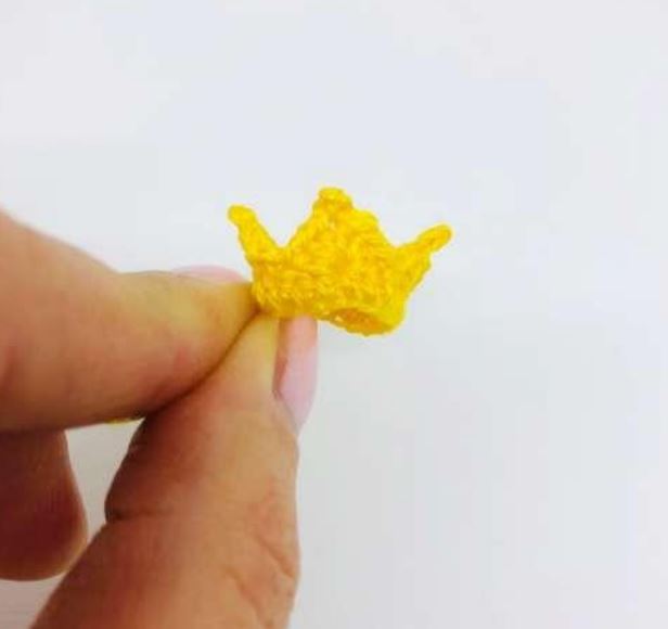 Tiny yellow crochet crown amigurumi held between fingers, detailed with points and textured stitches.
