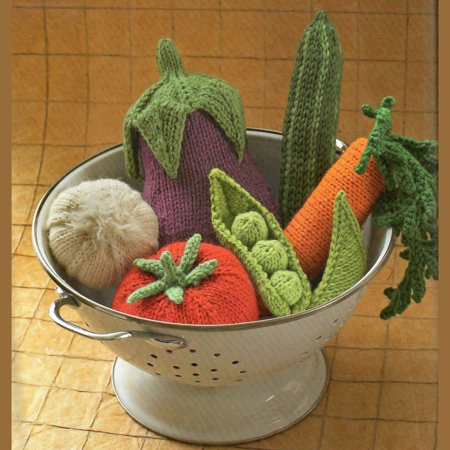 Knitted amigurumi vegetables including eggplant, carrot, tomato, cucumber, peas, and turnip in a metal colander.