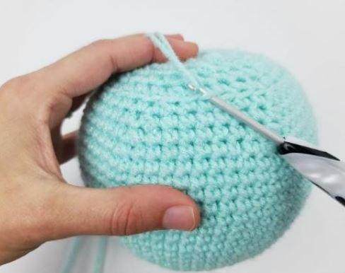 Hand holding light blue crocheted amigurumi ball with black and silver crochet hook inserted.